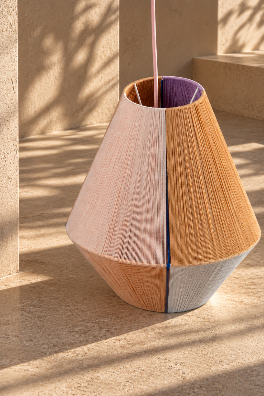 Close-up of the SYMI pendant lamp resting on a warm architectural stone surface, showing its hand-threaded texture and soft colour panels